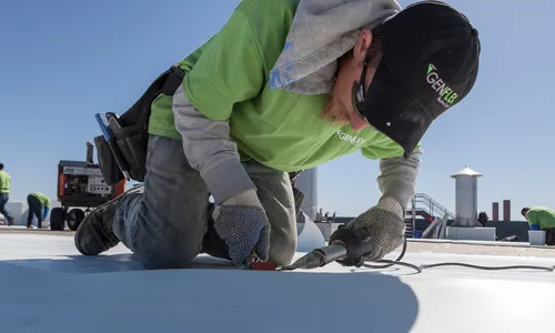 A worker in a green shirt and cap kneels on a rooftop, using a tool to install or repair roofing material under bright daylight. Other equipment and vents are visible in the background. | GenFlex