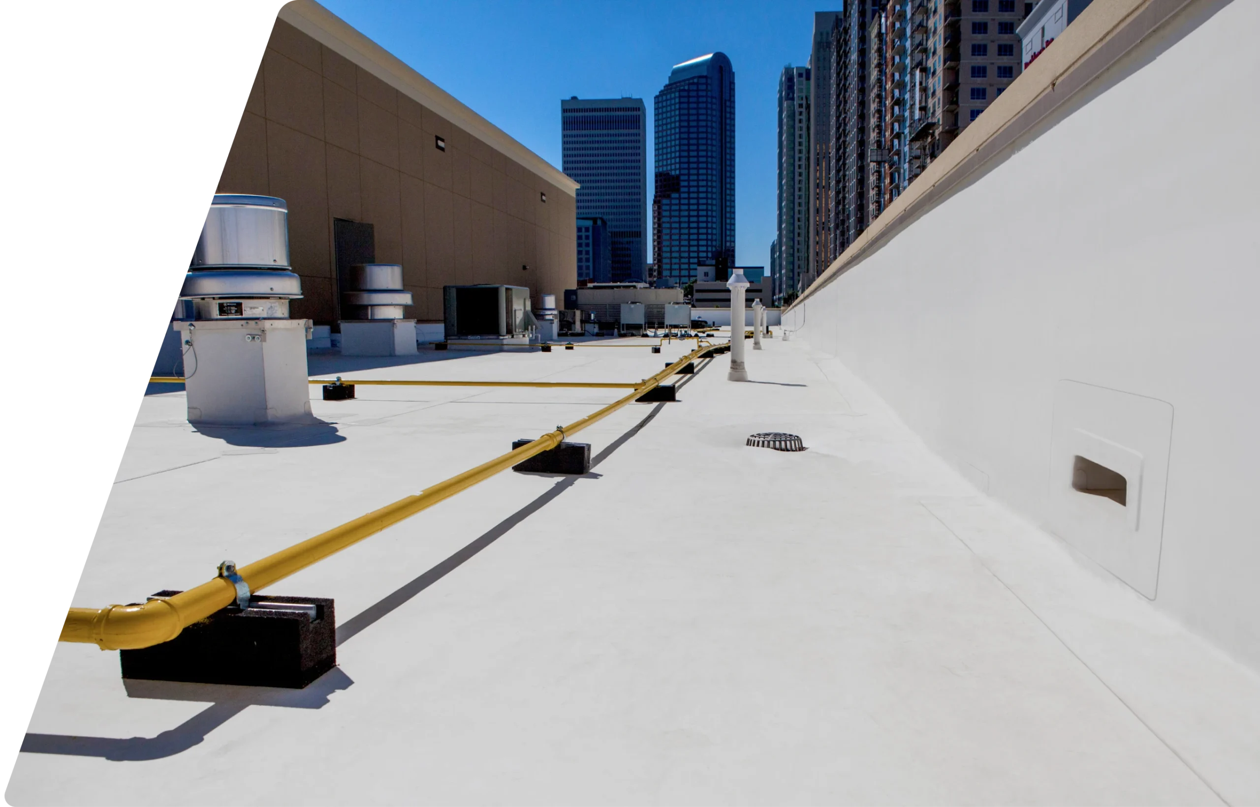 A white commercial rooftop with yellow gas pipes, ventilation units, and city skyscrapers in the background under a clear blue sky. The left side of the image is partially obscured by a dark vertical shadow. | GenFlex
