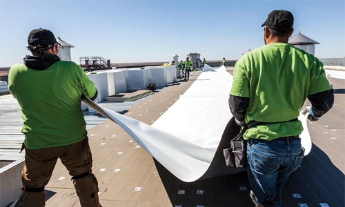 Two workers in green shirts unroll a large sheet of roofing material on a flat rooftop, with more workers in the background also participating in the installation under a clear sky. | GenFlex