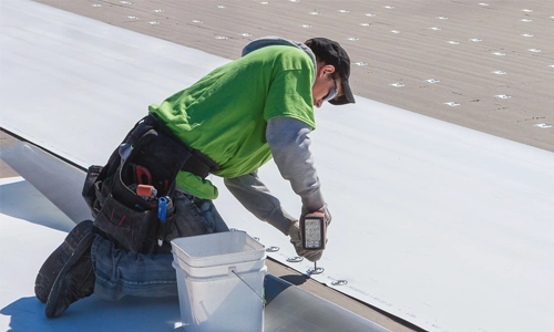 A worker in a green shirt and cap kneels on a roof, using a power tool to secure a white roofing material. He has a tool belt and a white bucket nearby. | GenFlex